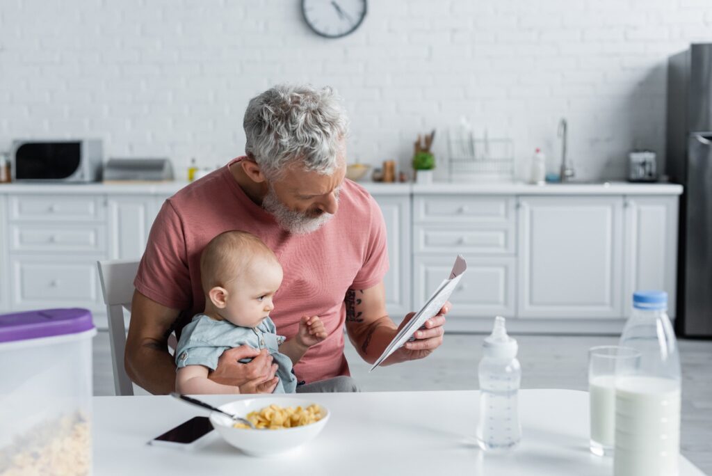 Mature Man Reading News Near Baby Daughter And Breakfast In Kitchen